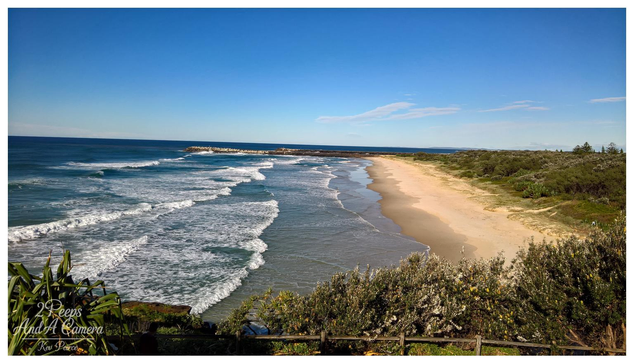 A sweeping, elevated view of a sandy beach on the Northern Rivers coast of New South Wales (NSW), south of Byron Bay. 

Powerful white waves roll in from the deep blue ocean, meeting the golden sand. Coastal scrub and a wooden fence line the foreground, with a distant rock breakwater visible.