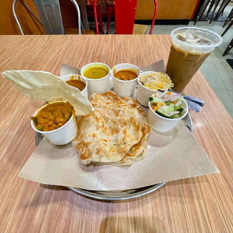 A platter featuring two pieces of prata, accompanied by four small cups of curry dishes and one cup of rice. A cup of veggies is present, along with an iced coffee drink in a large cup on the right. These are all placed on a wooden table.