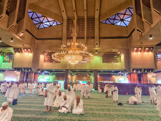 A wide interior photograph of a large mosque hall filled with dozens of Muslim men. Almost all the men are wearing the white, two-piece Ihram pilgrimage garments. They are scattered across a green patterned carpet; some are standing and walking, while others are sitting or performing prayers (kneeling and prostrating). The hall features high, brutalist-style concrete architecture with geometric skylights and a massive, ornate crystal chandelier hanging in the center.