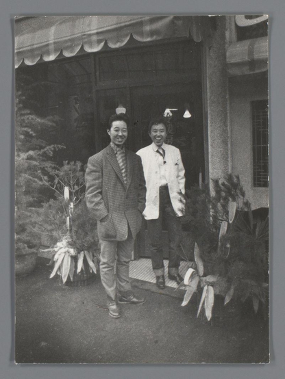 The image is a black and white photograph featuring two men standing in front of what appears to be the entrance of an establishment. The man on the left, wearing glasses, has his hands tucked into his pockets and sports a suit with a patterned tie; he's smiling broadly at the camera. Beside him stands another individual dressed in a light-colored jacket over a white shirt, also sporting a wide smile as they gaze directly ahead.

Behind them is an entrance marked by wooden beams overhead adorned with what seems to be fabric or paper decorations hanging on both sides. Flanking this central scene are potted plants and decorative elements that add character to the setting. The overall ambience suggests a casual yet festive occasion, possibly related to local customs or celebrations in Osaka.

Additional information about this image is found under 'Twee mannen voor een café in Osaka', Ed van der Elsken, 1960 [source provided], which points towards Dutch text and seems unrelated contextually. This indicates the photograph might be part of a larger collection or series by photographer Edward van der Elsken documenting life in Japan during that era.