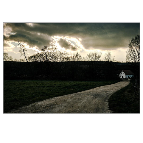 On the right side of the picture, a rural farm can be seen in the distance. The road leading there starts in the foreground of the picture. A white cobblestone road. The picture was taken at dusk, the colors are already faded, dark, stormy clouds cover the sky and the slowly setting sun.