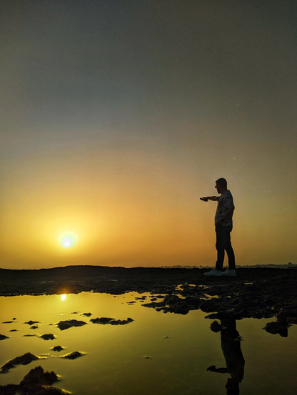 Image of a sunset, with a man visible on the right pointing at the sun. The shot was taken in a rocky beach and there’s a reflection of the scene in a small ocean water puddle