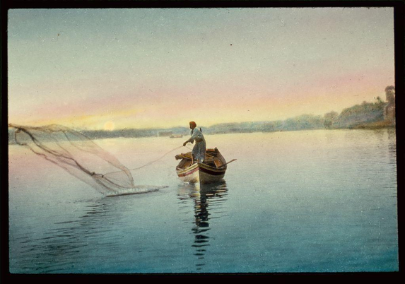 The image depicts a serene scene of a person fishing on a calm body of water. The person is standing in a small wooden boat, which appears to be a traditional fishing boat, and is holding a fishing net that is partially extended into the water. The individual is wearing a dark-colored outfit, possibly a smock or a similar type of clothing, suitable for fishing activities. The water is still, reflecting the soft colors of the sky, which transitions from a light blue at the top to a warm, pinkish hue near the horizon, suggesting either sunrise or sunset. In the background, there are faint outlines of land with trees and possibly some structures, adding depth to the scene. The overall atmosphere of the image is peaceful and tranquil, evoking a sense of quiet solitude and connection with nature.
