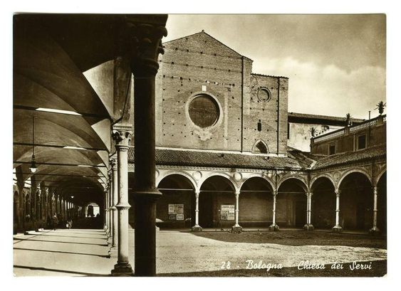 This image appears to be a historical black-and-white photograph of a religious or institutional building, specifically identified as "Chiesa dei Servi" (Church of the Servants) in Bologna, Italy. The structure is characterized by its brick construction and a large circular window in the central facade, which is a notable feature of the church. The roof is tiled and has a simple, sloped design. 

In the foreground, there is a covered walkway with arches supported by columns, creating a colonnade. The columns are evenly spaced and have a classical design, suggesting a Renaissance or Gothic architectural style. The ground is paved, and there are some people visible in the distance, giving a sense of scale to the scene. The sky above is partly cloudy, indicating that the photo was taken during the daytime. 

The image has a vintage quality, with a sepia tone and a slightly faded appearance, which suggests that it is an older photograph, possibly from the early to mid-20th century. The bottom right corner of the image has text that reads "28. Bologna Chiesa dei Servi," which confirms the location and subject of the photograph.