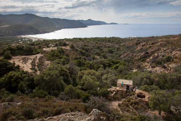 A scenic landscape featuring a coastal view with mountains in the background. There is a small building near the foreground, surrounded by greenery and rocky terrain. People are visible near the structure.