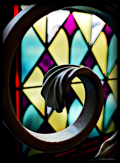 École d'art de Riom : fer forgé formant une volute en forme de fleur devant un vitrail aux formes de losanges jaune, vert, rose.