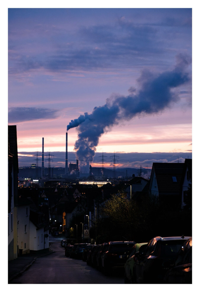 Foto im Hochformat. Blaue Stunde, kurz nach Sonnenuntergang. Blick entlang einer abschüssigen Straße. Rechts parkt ein Auto nach dem anderen, links sind Häuser. Richtung Horizont ist ein großes Kraftwerk im Tal. Aus mehreren Schornsteinen steigt Rauch auf, der in einer dicken Wolke vom wind nach rechts getragen wird. Der Himmel am Horizont ist teils bewölkt und rötlich-orange. Darüber, am bläulichen Himmel sind weitere Wolken.
