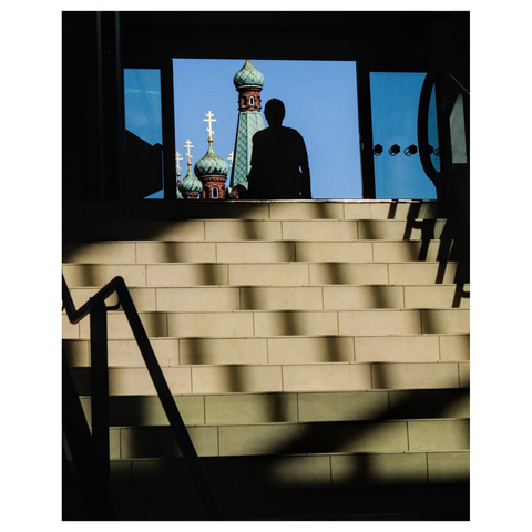 This image shows a silhouette of a person walking at the top of a well-lit staircase with white /beige steps. Through a large window or opening in front of the person, colorful orthodox church domes with golden crosses are visible against a clear blue sky. The domes are ornate, with green and gold details. The staircase is indoors, and the lighting creates strong shadows on the steps. A metal handrail is visible on the left side of the staircase. (draft Mistral.ai, edited by author)