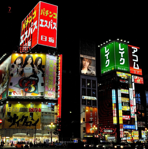 Photo couleur de nuit d'un quartier japonais de Tokyo.
Plusieurs immeubles éclairés sont pris en photo. Sur ceux-ci des mots en japonais mais aussi des images qui indiquent les commerces occupants les immeubles. En bas de la photo, la rue avec des voitures les phares allumés et des personnes  qui marchent sur le trottoir.