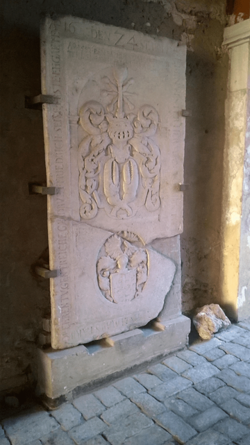 Photo of a broken stone door, with decorative design (lower part of a very fancy coat of arms and upper part looks like a flowery pot coat of arms), with Latin writing on the sides of the door, held to the wall by metal clamps.