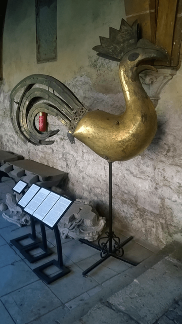 Photo of a large metal rooster - colored gold - but looks a bit rusted and not as shiny, leaned against an old stone wall, with pieces of stone decorations laid on the ground.
