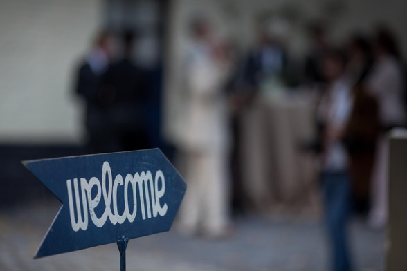 A “welcome” sign in the foreground with a blurry background showing a group of people.