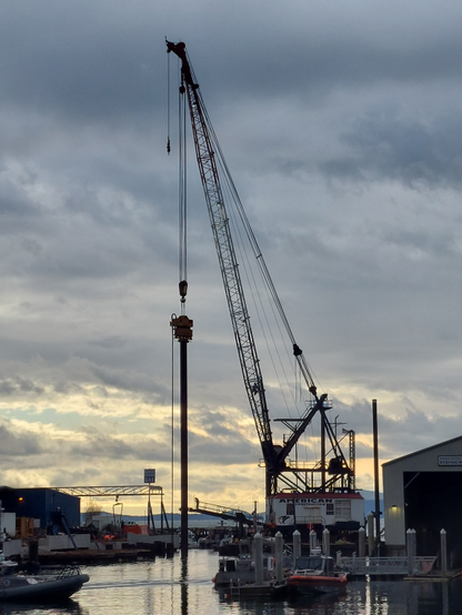 a scene at harbor, with some construction rig type mining equipment reaching from high into the sky down into the water, illuminated by a false sunset - the sun is not quite on the horizon yet, but the cloud cover breaks only along the horizon, giving the soft illusion that that's where the only sunlight is