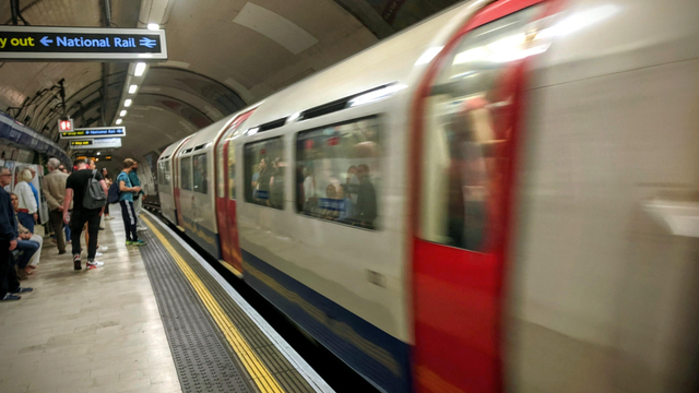 A blurred, long exposure shot capturing a London Underground train as it pulls rapidly into a station tunnel. The platform on the left is in focus, showing several passengers waiting or walking, with "Way out \rightarrow National Rail" signs visible overhead.