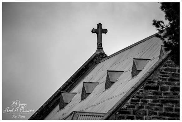 Black and white photograph showing a dramatic, close up, upward angled view of a church roof.

The roof is corrugated iron with three pointed dormers, topped by a stone cross silhouette against a heavily clouded sky. The texture of the stone wall is visible below the roofline.