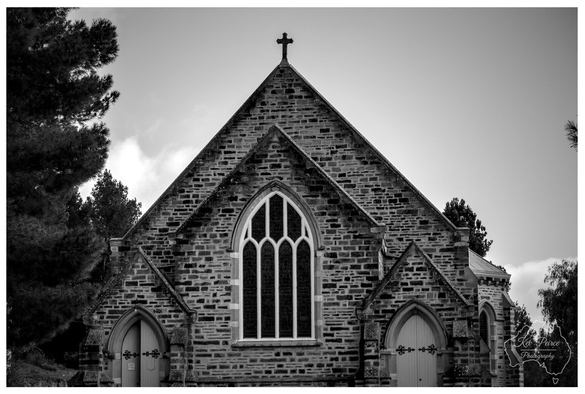 Black and white photograph capturing a frontal, symmetrical view of a historic stone church's facade.  Features include a large Gothic style arched window with white tracery, two smaller side entrances, and a cross atop the main gable. Dark trees frame the structure on the left against a bright sky.