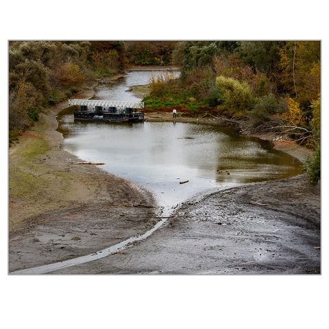 The picture shows a drought-stricken bay in the fall. A pontoon bridge is stuck in the bay bed. A man in a white striped shirt stands next to it, looking out at the water.