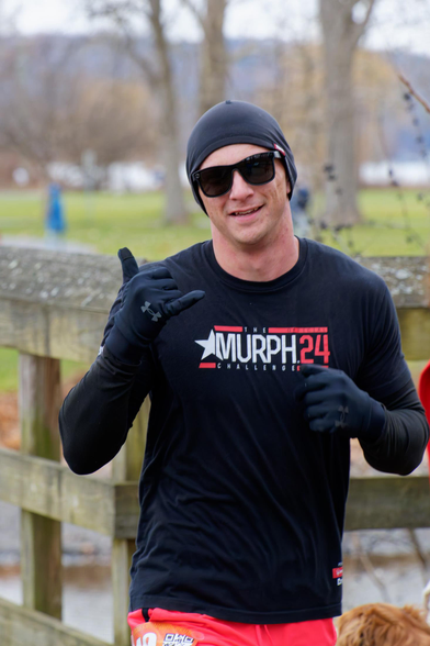 Man in black shirt that says MURPH 24 in red and white makes a gesture with a gloved hand with the backdrop of a pretty park by a lake