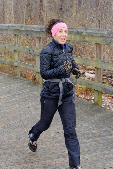 Woman in black coat with wide pink headband runs down a wood bridge.