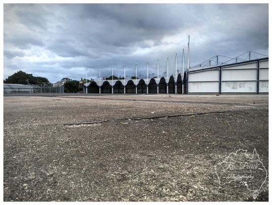 A low angle, wide shot of a large, desolate, cracked asphalt or concrete expanse leading toward the industrial sheds of a former rail yard under a heavily overcast, moody sky.

In the mid ground, a long, curved row of shed entrances with a distinctive, pointed, scalloped roofline provides a strong geometric focal point, flanked by a large white building on the right.