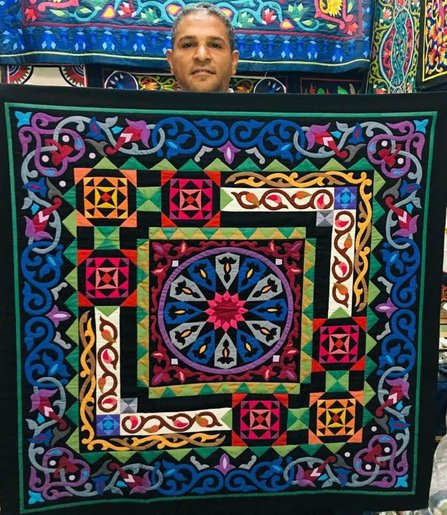 Photo of one of the last Egyptian khayamiya makers holding up an intricately patterned, brightly colored example of his work. Behind him are more of his artworks.