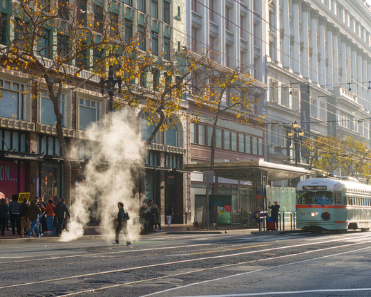 A woman walks through steam rising from a grille in the street, with a vintage streetcar in the background