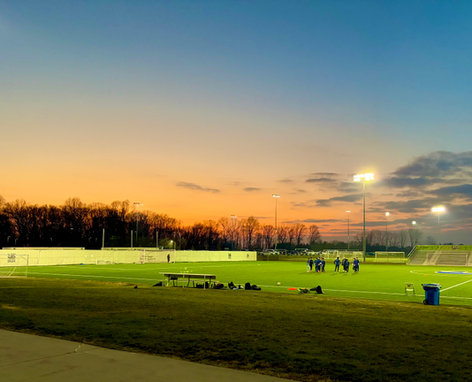 A brightly lit soccer field glows with vibrant green turf under lights. Above it, the sky transitions from a warm orange near the horizon to blue overhead. On the field, a group of youth soccer players wearing navy uniforms gather together on the turf.