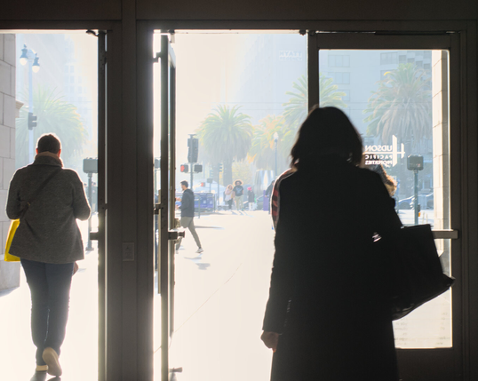 A woman in a winter coat is silhouetted against bright sunlight streaming through an open door