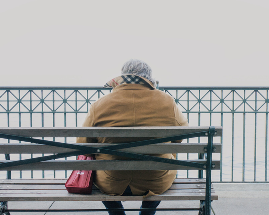 A man in a winter coat with the collar upturned against the cold, sits on a bench looking out on the foggy ocean