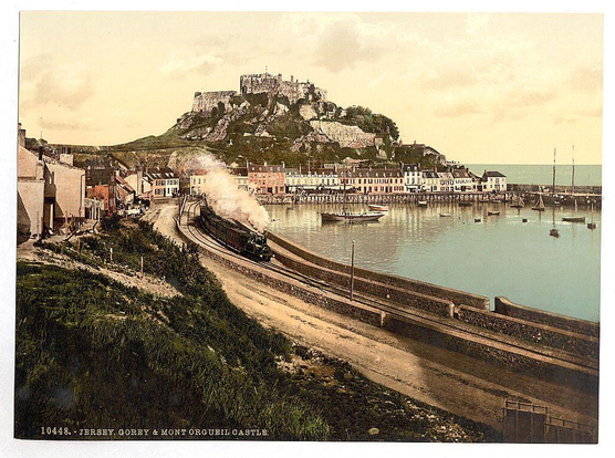 The image is a historical photograph showcasing a coastal town with a prominent castle perched atop a hill in the background. The scene is vibrant, with the castle standing out due to its stone structure and the surrounding greenery. The town is nestled along the shoreline, featuring a mix of buildings with pitched roofs and chimneys, suggesting a European architectural style. A train, emitting a plume of steam, is visible on a railway track running parallel to the shoreline, adding a sense of movement and activity to the scene. The harbor area in the foreground is dotted with small boats, indicating a bustling port. The sky is partly cloudy, with soft, diffused lighting, giving the image a serene and timeless quality. The text at the bottom of the image identifies the location as "Jersey, Gorey & Mont Orgueil Castle."