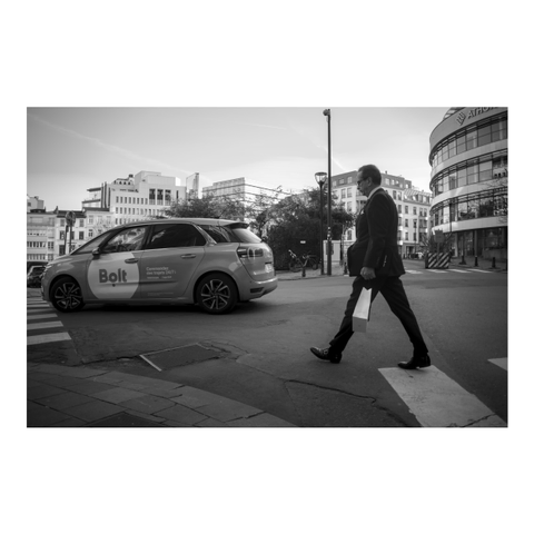 A black and white photo of a man in a dark suit crossing a street in a city. He is holding a small white paper bag and a dark, small briefcase in his hand next to his body. The photo captures him in the middle of a step. A car with the label "bolt" is driving in front of him.