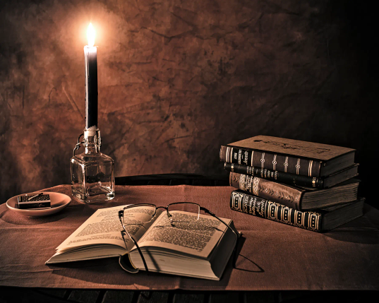 Auf einem Tisch liegt ein aufgeschlagenes Buch, eine Brille ist auf den Seiten abgelegt. 
Dahinter auf dem Tisch links eine brennende Kerze in einer Flasche, daneben eine kleine Schale mit einer Streichholzschachtel. Ein abgebranntes Streichholz liegt oben drauf. Rechts liegen drei weitere, sehr alte Bücher gestapelt. 