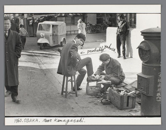 The image depicts a street scene in what appears to be Osaka, Japan from 1960. In the foreground, there's an individual sitting on a stool with another person crouched beside them working on polishing shoes using a cloth and bottle of polish. They are surrounded by various objects such as a suitcase for storage, bottles presumably containing cleaning solutions or shoe polish, and other miscellaneous items in crates that suggest they might be selling their goods.

In the background, several people can be seen going about their business—walking down the street, standing against walls, and engaging with others. The setting appears to be an urban environment with a mix of traditional Japanese elements like the postbox labeled "POST" and modern features such as parked vehicles including what looks like early models of vans or cars.

The overall atmosphere captures daily life on a bustling city street during that time period in Japan's history, showcasing both personal interactions (like the shoe shining) and broader societal context through visible activity. The scene is devoid of color due to being monochromatic which emphasizes textures, shapes, and contrasts more than any specific hue or saturation.

The image serves as an important historical document reflecting a slice of life during that era in Osaka.