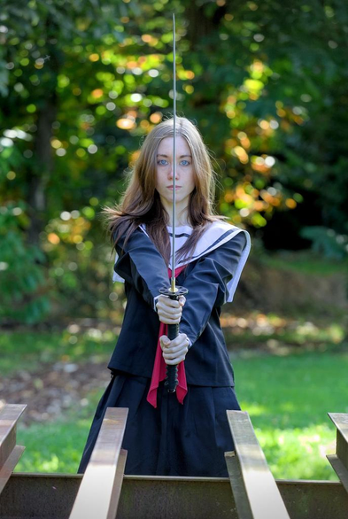 Photo format portrait d’une jeune femme en tenue d’écolière japonaise bleu marine, dotée d’un ruban rouge et d’un col blanc, tenant un katana droit devant elle, partageant son visage en deux. Elle regarde droit devant elle vers la caméra d’un air glacial, renforcé par la couleur remarquable de ses yeux bleu acier. Au premier plan, un bout de structure en métal rouillé avec des poutres en acier, dans l’axe de perspective. Le fond est flou et composé de verdure avec des tâches de soleil dedans
