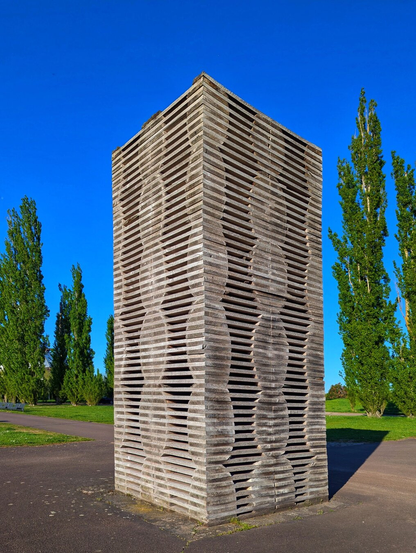 This image depicts a tall, rectangular outdoor sculpture crafted from horizontally stacked layers of sawed oak wood. The structure features an intricate design with curved, cut-out sections that form a visually engaging pattern. The oak trunks have been precisely sawed and reassembled, giving the sculpture a distinctive, textured appearance. The sculpture stands on a paved surface, set against a backdrop of a grassy area and tall, slender trees with vibrant green foliage. Above, the sky is a clear, bright blue, suggesting a sunny day.