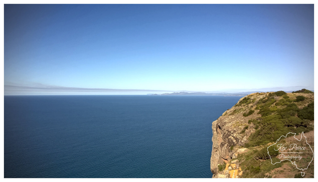 A wide angle view from a rocky, scrub covered cliff on the right, looking out over a deep blue ocean that extends to the horizon.  The sky is a clear, lighter blue, meeting the sea with a subtle distant landmass visible.  The photo is signed 'Kev Peirce'.