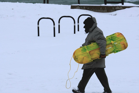 A sledder carries a yellow sled while wrapped up in a heavy jacket and face covering. There is a bicycle rack in the background. The scene is snow covered.
