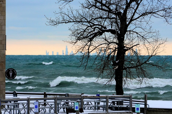 A view of the Chicago skyline with the edge of the pavilion on the left side. There is also a patio surrounded with a railing, three disabled parking signs, a barren tree, and waves crashing into the Indiana Dunes with white caps.