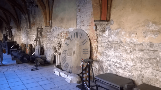 Photo taken from inside of an old church side building hallway, with an old clock face, dusty and not-cleaned-in-ages-type dirty, leaned against the old brick wall, with stone chairs and stone and metal decorations, placed on the floor.