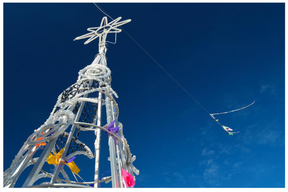Querfomat: links wächst ein pures weisses Weihnachtsbaumdrahtgestell mit Lichtern und bunten Wimpeln in den Himmel, ein Kabel schwebt mit weiteren Plastikwimpeln noch etwas höher in die klare Winterluft. Knallblauer Hintegrund.