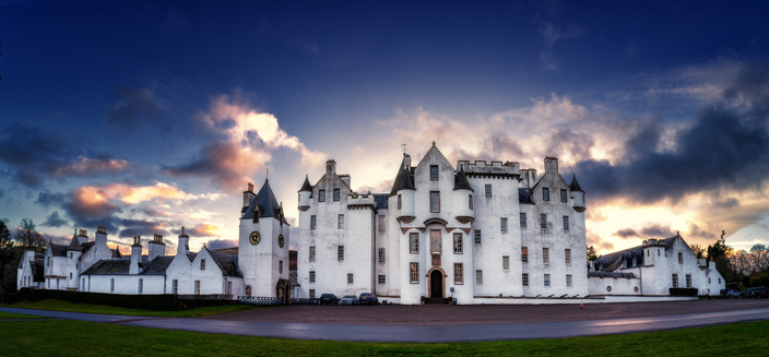 Der Panoramablick zeigt Blair Castle, ein markantes weisses historisches Gebaeude, vor einem dramatischen, farbenfrohen Himmel bei Sonnenuntergang. Die Komposition hebt die architektonischen Details des Schlosses, einschliesslich seiner Tuerme und Fenster, hervor und kontrastiert sie mit der umgebenden gruenen Landschaft. Das Zusammenspiel von beleuchteten Wolken und der hellen Fassade des Schlosses schafft eine eindrucksvolle visuelle Darstellung.