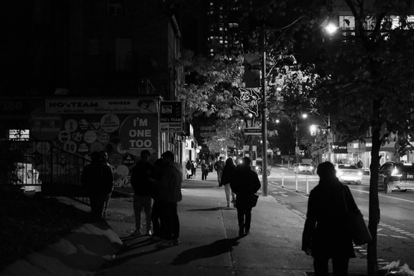 Night, looking toward Church Street. The lights of the intersection dominate the upper right background of the frame, though the rest is deep in shadow. We see silhouettes of the pedestrians on the sidewalk and just a hint of the murals on the walls of the buildings on the left. 