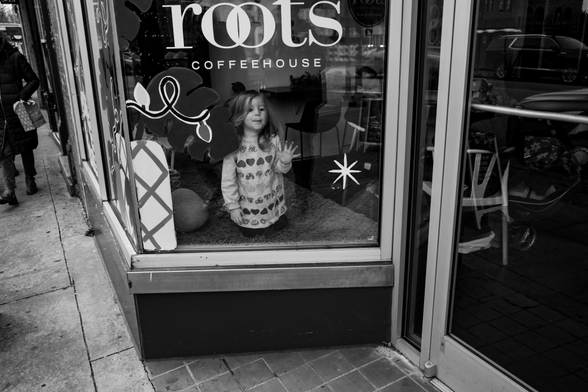 A black and white photo from outside a coffee shop.  There is a little girl inside on her knees pressing her face against the window.