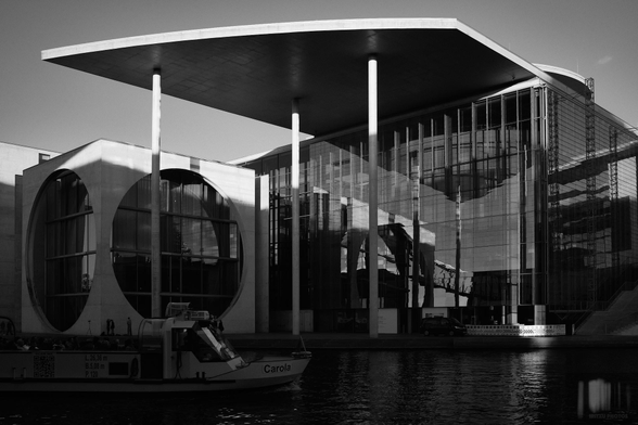 Black-and-white photograph from the government district overlooking the River Spree, where a ship is just entering the frame, with the Marie-Elisabeth-Lüders-Haus on the opposite bank in Berlin, Germany, in the background.