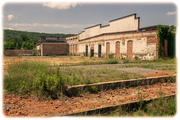 An abandoned industrial complex with deteriorating red brick and white-painted masonry buildings. The structures show significant weathering with exposed brick, peeling paint, and bricked-up arched windows and doorways. In the foreground, a crumbling concrete loading dock or platform is littered with broken red bricks and debris, with weeds growing through the cracks. The surrounding area is overgrown with wild grasses and sparse vegetation on bare, compacted earth. Green wooded hills rise in the background under an overcast sky with scattered clouds. The image has a vintage-style vignette border effect. Trees are visible on the left side and behind the buildings, emphasizing the site's abandonment and return to nature.