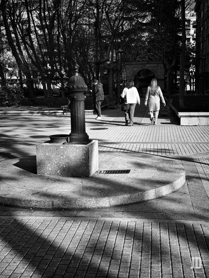 Fotografía en blanco y negro de un parque urbano soleado con fuertes contrastes. En primer plano, a la izquierda, se alza una fuente de agua antigua de metal oscuro sobre una base de piedra rectangular, rodeada por una plataforma circular de granito. El suelo, pavimentado con baldosas, está cruzado por largas y nítidas sombras proyectadas por los árboles. Tres personas caminan por el sendero principal, de espaldas al espectador: un hombre con chaqueta oscura a la izquierda y dos mujeres en el centro, una con una sudadera clara y bolso, y la otra con un abrigo más largo. El fondo está dominado por densos árboles oscuros y sin hojas, y entre ellos se vislumbra un arco de piedra o mampostería, con parte de un edificio a la derecha.