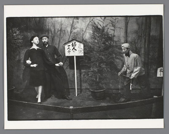 The image is a black and white photograph depicting three individuals on what appears to be an outdoor stage with a backdrop of trees. On the left, there are two people standing close together; one person seems female wearing dark clothing including a long-sleeve top and skirt, while the other individual's gender cannot be determined due to their attire which includes overalls and a cap suggesting male characteristics. The central focus is on another figure with an appearance resembling that of a man in traditional Japanese attire, possibly from historical or cultural context given his simple, practical outfit consisting of a jacket, trousers, hat, boots, and holding what looks like a fishing rod. In the background stands a signpost indicating "one step to woman's house" which may suggest narrative significance within this performance scenario. There is also another smaller placard with Japanese writing on it located closer to the right side figure. The setting seems staged for an audience or as part of cultural exhibition, and there are props such as buckets placed near the figures adding context to their roles in what could be a theatrical production or street performance piece relating to traditional customs or social commentary within Japan during that era.