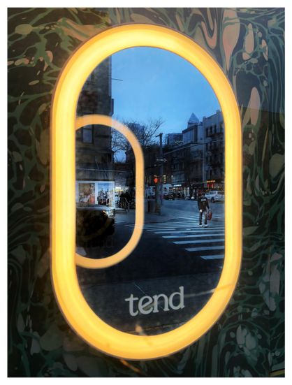 Photo, taken at a slight angle, of a store window set into a wall with a swirling green pattern that suggests leaves and flowers. The window is capsule-shaped with a rounded top and bottom and has a wide border of luminous yellow light. Inside the window, off-center to the left, is another capsule outline that appears to float unsupported in the air; it’s unclear if this is a reflection or some other optical effect. The impression is of two glowing rings, the inner one receding into the distance. The window glass reflects a city scene outside: a crosswalk and low-rise buildings under a blue evening sky. A pedestrian crosses the street near the right edge of the outer ring; the inner ring frames a car’s headlights and oversized ads with images of models. At the window’s curved bottom is the word “tend” in an elegant lowercase font.