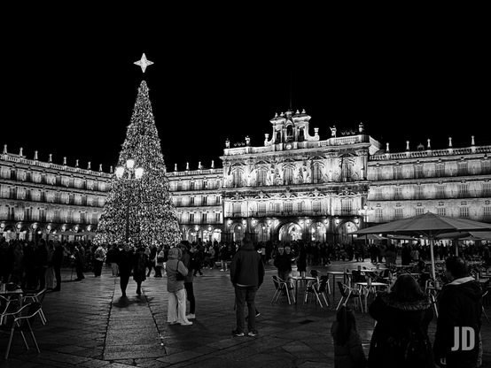 Fotografía en blanco y negro captura una escena nocturna festiva en la Plaza Mayor de Salamanca, España.
Los elementos principales incluyen:
• Árbol de Navidad: Una gran estructura cónica profusamente iluminada, coronada por una estrella brillante, actúa como punto focal en el centro de la plaza.
• Arquitectura Histórica: Al fondo se aprecia la majestuosa fachada barroca del Ayuntamiento y los edificios con soportales que rodean el recinto, todos destacados por la iluminación nocturna.
• Multitud y Ambiente: Numerosas personas pasean y se reúnen alrededor del árbol, creando una atmósfera vibrante y concurrida.
• Primer Plano: Se observan mesas y sillas de las terrazas de los cafés, dispuestas para los visitantes en este espacio emblemático.