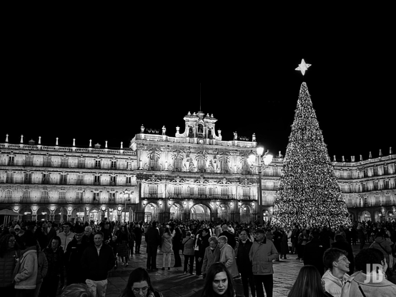 Serie de imágenes en blanco y negro captura diferentes perspectivas nocturnas de la Plaza Mayor de Salamanca, España, durante la época navideña.
Los elementos clave que se aprecian son:
• Punto Focal Navideño: Un gran árbol de Navidad cónico y profusamente iluminado, coronado por una estrella brillante, se sitúa en el centro de la plaza.
• Arquitectura Barroca: Las imágenes muestran la fachada del Ayuntamiento y los edificios con soportales que rodean el recinto, destacados por una iluminación monumental.
• Vida Urbana: Se observa una gran afluencia de personas paseando y disfrutando de la atmósfera festiva.
• Mobiliario de Terrazas: En los primeros planos se distinguen las mesas, sillas y sombrillas de los cafés de la plaza, integrándose en la escena cotidiana.